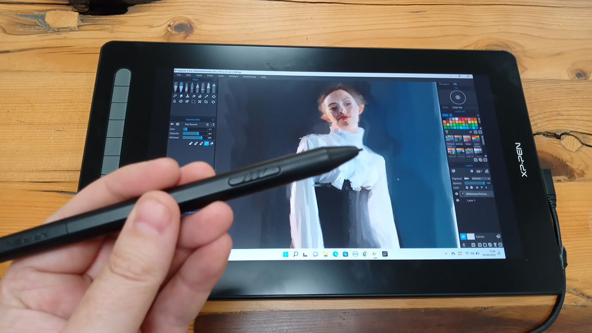 A photo of a hand holding a stylus in front of the XP-Pen Artist 16 (2nd gen) drawing tablet.