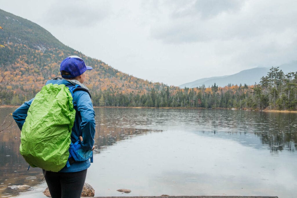 Female hiker looking out over lake in New Hampshire in the fall wearing backpack with rain cover